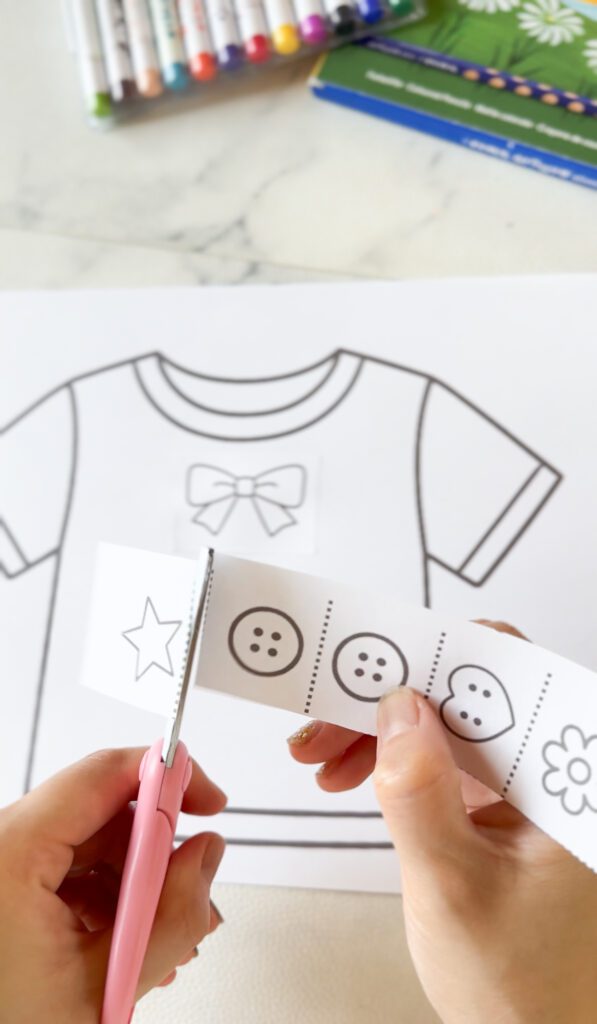 Tシャツが印刷されたプリントに、図形が描かれた紙を子ども用ハサミで切ってい、Tシャツに貼ろうとしている様子。雨の日のはさみ遊びの無料知育プリント教材